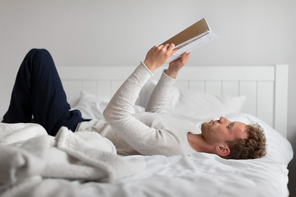 Comment s'occuper quand on est immobilisé ? 1 Personne immobilisée dans un lit avec une jambe dans le plâtre, lisant un livre avec un plateau-repas à côté.