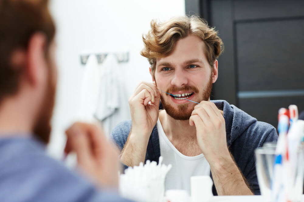 Homme se regardant dans le miroir en passant du fil dentaire, gêné par une sensation persistante entre deux dents.