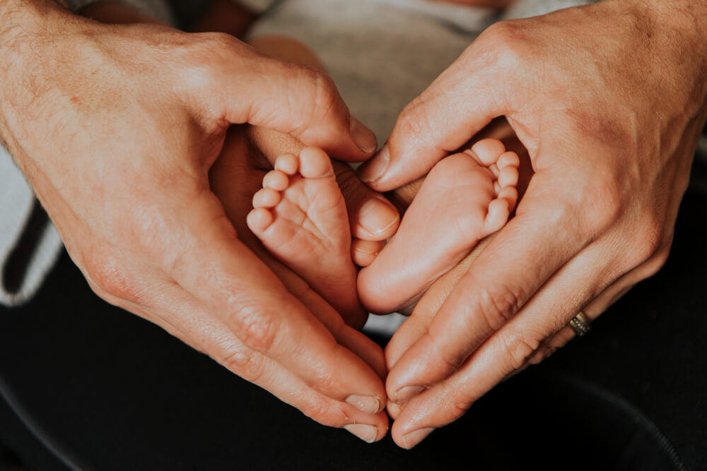 Naissance : les 10 premières choses à faire dans le premier mois (sans se noyer) 3 Gros plan sur les mains d'un parent tenant délicatement les pieds d'un nouveau-né durant un moment de soin à la maison.