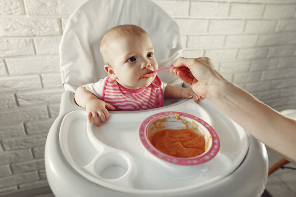 Bébé a mal au ventre depuis la diversification : solutions et explications simples 2 Un adorable bébé assis dans sa chaise haute qui goûte sa première purée de carottes avec une cuillère en silicone.
