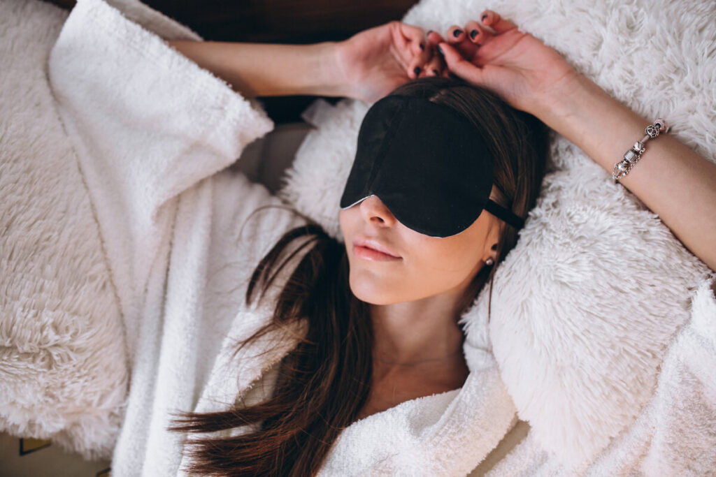 Les bienfaits d’un masque anti-migraine 2 Une personne allongée dans le noir portant un masque compressif en gel froid sur les yeux pour soulager une crise de migraine.